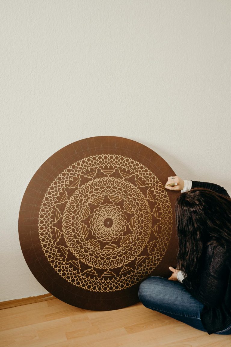 A woman sitting indoors next to an intricately detailed round mandala art piece, adding a warm and artistic touch.